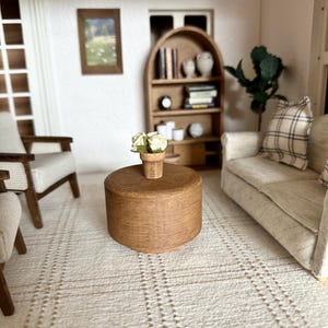 Op de afbeelding: Miniatuur woonkamer sc&egrave;ne met een ronde, bruine salontafel met een kleine pot met witte bloemen. De kamer omvat een beige bank met een geruit kussen, twee fauteuils, een boekenkast en een ingelijste foto. Het tapijt heeft een ruitpatroon.