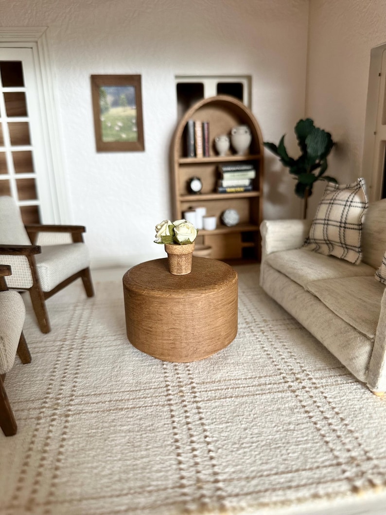 Op de afbeelding: Miniatuur woonkamer sc&egrave;ne met een ronde, bruine salontafel met een kleine pot met witte bloemen. De kamer omvat een beige bank met een geruit kussen, twee fauteuils, een boekenkast en een ingelijste foto. Het tapijt heeft een ruitpatroon.