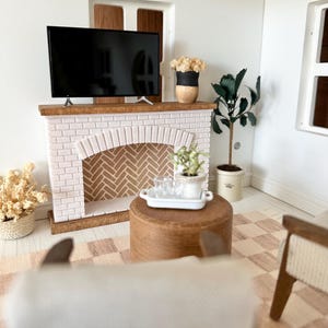 May include: Miniature living room scene with a white brick fireplace, a flat-screen TV, and a wooden coffee table. The room is decorated with plants, a patterned rug, and a neutral color palette. A small white tray holds glasses.