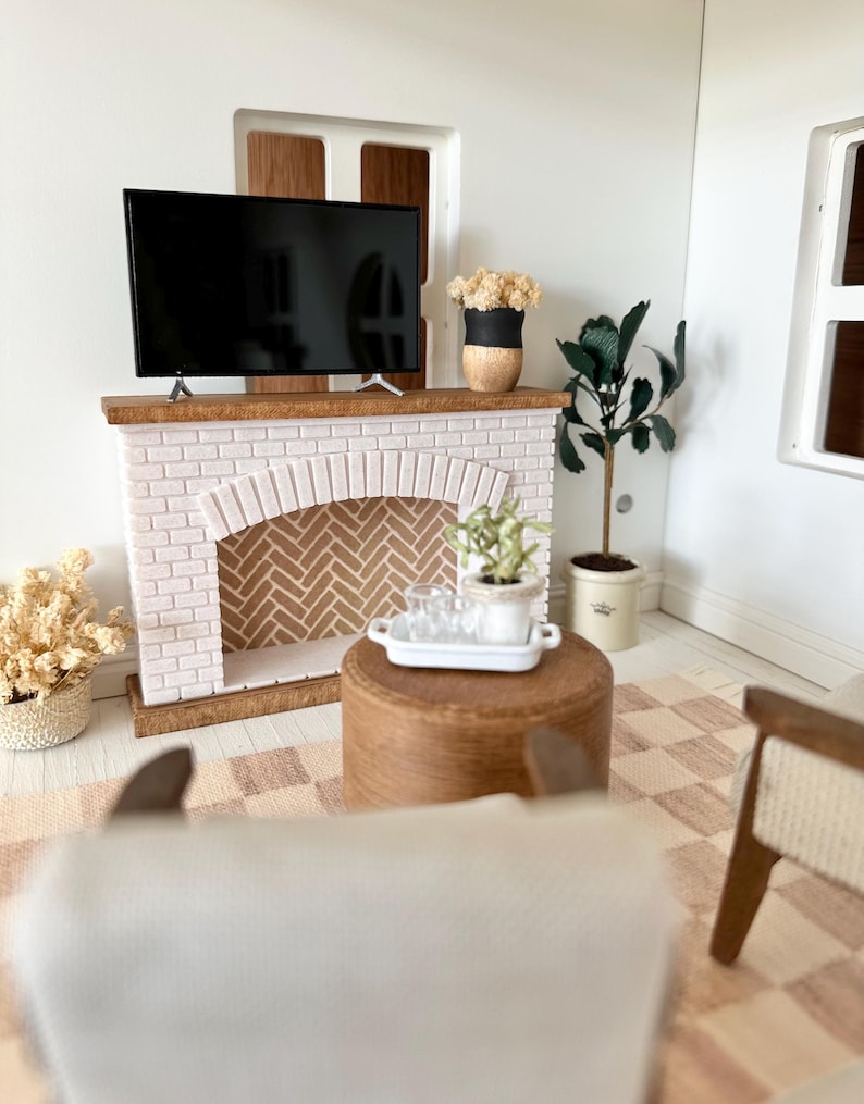 May include: Miniature living room scene with a white brick fireplace, a flat-screen TV, and a wooden coffee table. The room is decorated with plants, a patterned rug, and a neutral color palette. A small white tray holds glasses.