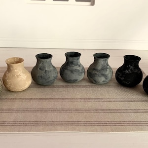 May include: Seven ceramic vases in various shades of grey and beige. The vases are arranged in a row on a striped rug.