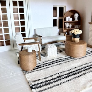May include: Miniature living room scene with a white sofa, armchair, and ottoman. A striped rug, two wooden side tables, and a bookshelf with decorative items complete the scene. A small vase with flowers sits on one of the tables.