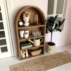 May include: A wooden arched shelf unit with four shelves, featuring a white bust, a small clock, a candle holder, glass cups, a watering can, books, and decorative white swans. The shelf unit is on a light wood floor with a brown woven rug in front of it.