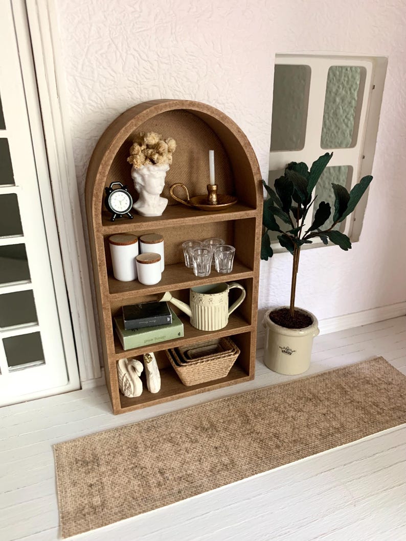 May include: A wooden arched shelf unit with four shelves, featuring a white bust, a small clock, a candle holder, glass cups, a watering can, books, and decorative white swans. The shelf unit is on a light wood floor with a brown woven rug in front of it.