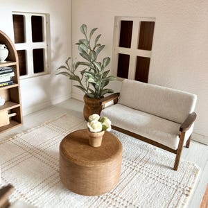 Op de afbeelding: Miniatuur woonkamersc&egrave;ne met een lichtgekleurde bank, een houten salontafel en een geweven vloerkleed. Een plant in een pot en een kleine vaas met bloemen voegen details toe. De kamer heeft twee ramen en een boekenplank.
