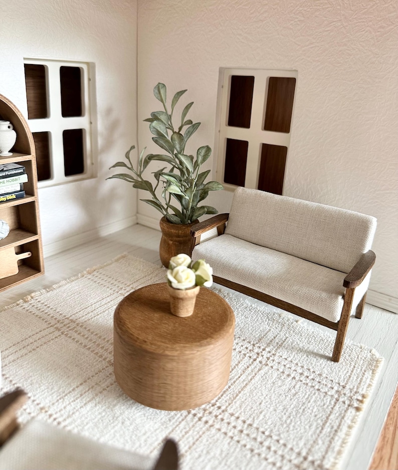 Op de afbeelding: Miniatuur woonkamersc&egrave;ne met een lichtgekleurde bank, een houten salontafel en een geweven vloerkleed. Een plant in een pot en een kleine vaas met bloemen voegen details toe. De kamer heeft twee ramen en een boekenplank.
