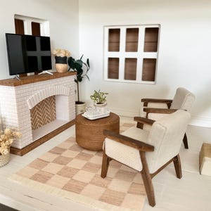 May include: Miniature living room scene with a white brick fireplace, a flat-screen TV, and a checkered rug. Two armchairs with light-colored upholstery and wooden frames face the fireplace. A round ottoman and decorative plants complete the scene.