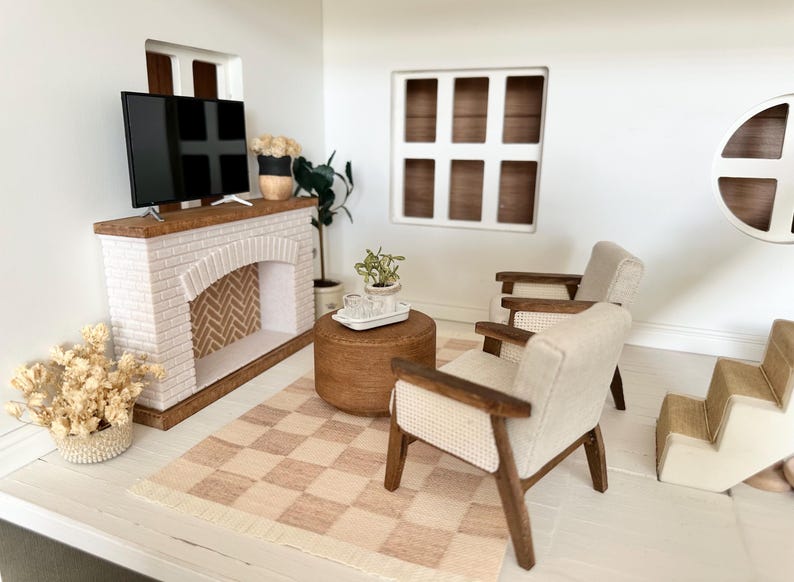 May include: Miniature living room scene with a white brick fireplace, a flat-screen TV, and a checkered rug. Two armchairs with light-colored upholstery and wooden frames face the fireplace. A round ottoman and decorative plants complete the scene.