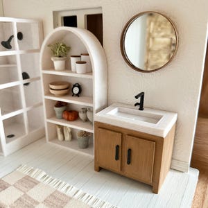 May include: Miniature bathroom scene with a wooden vanity, a white sink, and a black faucet. A round mirror hangs on the wall. A checkered rug and a white arched shelf with decorative items complete the scene. The color palette is neutral with wood tones.