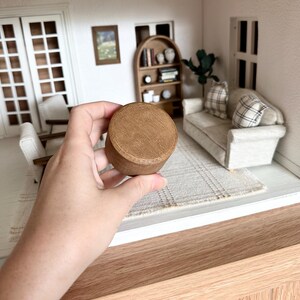 Op de afbeelding: Een kleine, ronde houten container met een deksel, vastgehouden in een hand. De container is lichtbruin van kleur en lijkt deel uit te maken van een poppenhuis of miniatuursc&egrave;ne. De achtergrond toont een poppenhuisinterieur met meubels.
