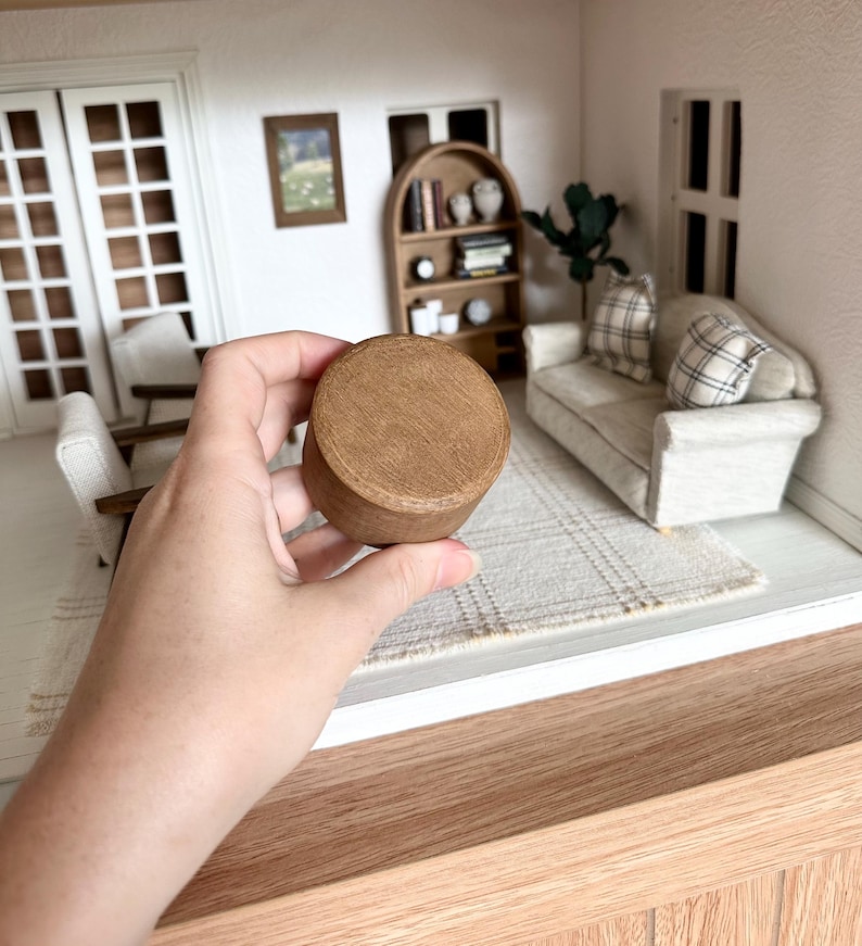 Op de afbeelding: Een kleine, ronde houten container met een deksel, vastgehouden in een hand. De container is lichtbruin van kleur en lijkt deel uit te maken van een poppenhuis of miniatuursc&egrave;ne. De achtergrond toont een poppenhuisinterieur met meubels.