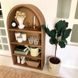 May include: A brown wooden arched shelf with four shelves, decorated with a white bust, a gold candle holder, a clock, jars, glasses, books, a watering can, white swan figurines, and a wicker basket. A potted green plant sits on the floor next to the shelf.