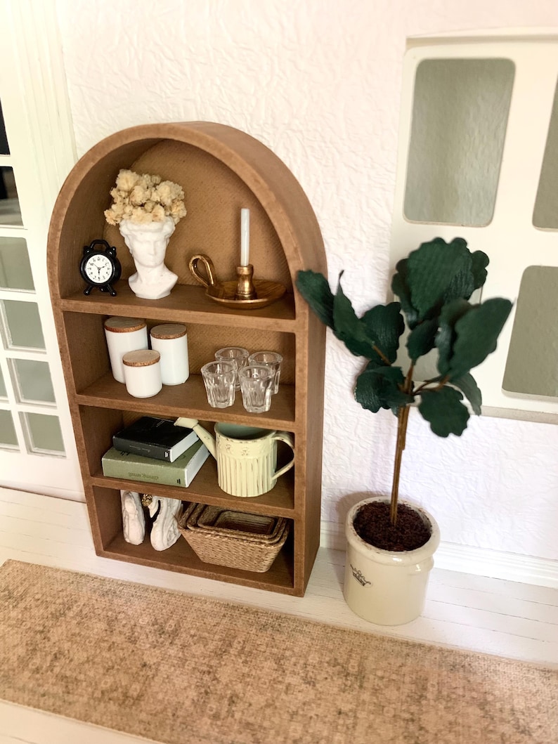 May include: A brown wooden arched shelf with four shelves, decorated with a white bust, a gold candle holder, a clock, jars, glasses, books, a watering can, white swan figurines, and a wicker basket. A potted green plant sits on the floor next to the shelf.