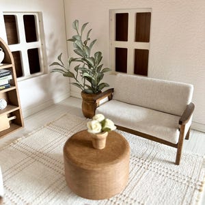 Op de afbeelding: Miniatuur poppenhuis woonkamer met een cr&egrave;mekleurige bank, houten salontafel en een geweven vloerkleed. Een potplant en een kleine vaas met witte bloemen voegen decoratieve accenten toe. De kamer heeft twee ramen en een boekenplank.