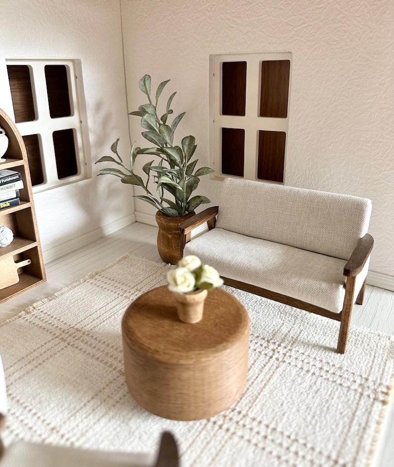 Op de afbeelding: Miniatuur poppenhuis woonkamer met een cr&egrave;mekleurige bank, houten salontafel en een geweven vloerkleed. Een potplant en een kleine vaas met witte bloemen voegen decoratieve accenten toe. De kamer heeft twee ramen en een boekenplank.