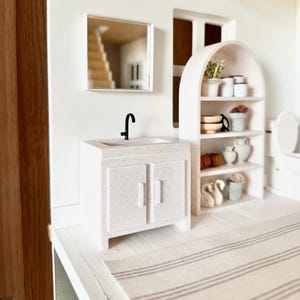 May include: A miniature bathroom vanity with a speckled off-white finish, a black faucet, and two cabinet doors. Beside it stands an arched shelving unit containing small decorative items, including white pots, bowls, and swan figurines.