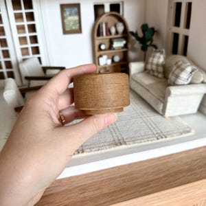 Op de afbeelding: Een miniatuur houten bijzettafel met een cilindrische vorm. De tafel is lichtbruin van kleur en heeft een textuur. De tafel is ontworpen voor een poppenhuis of miniatuursetting.
