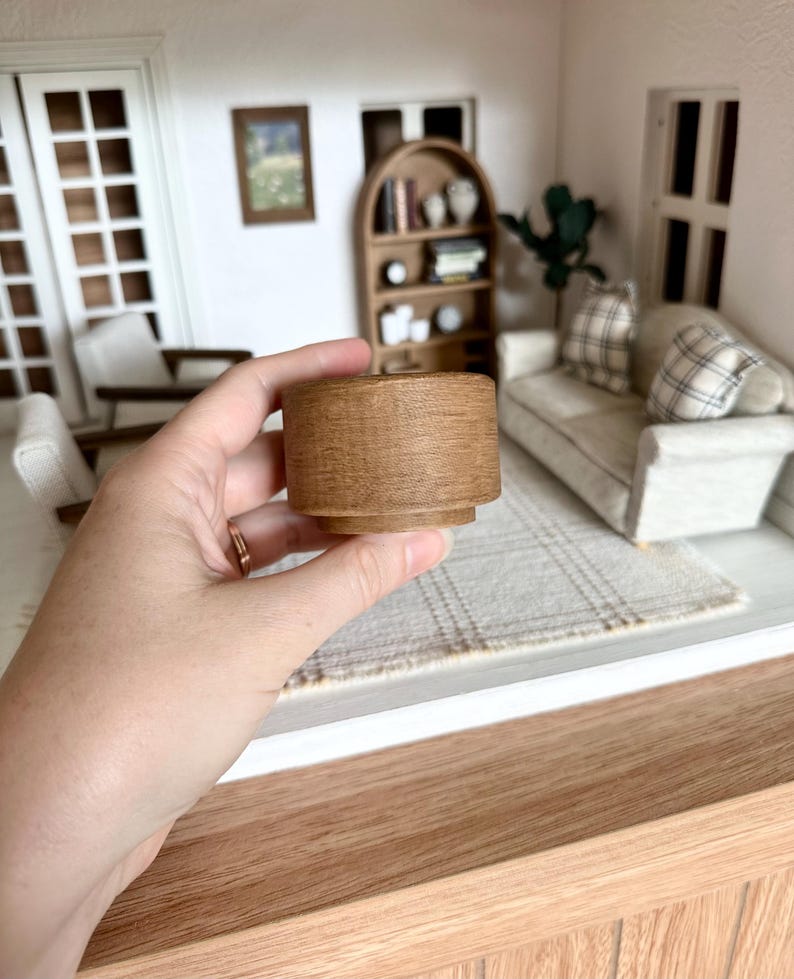 Op de afbeelding: Een miniatuur houten bijzettafel met een cilindrische vorm. De tafel is lichtbruin van kleur en heeft een textuur. De tafel is ontworpen voor een poppenhuis of miniatuursetting.