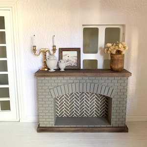 May include: A miniature fireplace with a gray brick facade and a herringbone pattern inside. The fireplace has a wooden mantel with two white ceramic vases and a gold candle holder. A framed picture and a vase with dried flowers are on the mantel.