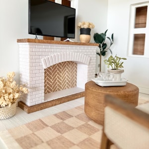 May include: Miniature living room scene with a white brick fireplace, a flat-screen TV, and a wooden coffee table. The fireplace has a herringbone pattern inside. A checkered rug and decorative plants complete the cozy interior.