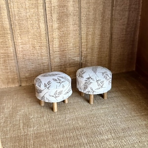 May include: Two miniature round stools featuring a cream-colored fabric upholstery with a dark brown botanical print of wheat stalks and leaves. Each stool is supported by four small, light-colored wooden legs, providing a stable base for the seat.