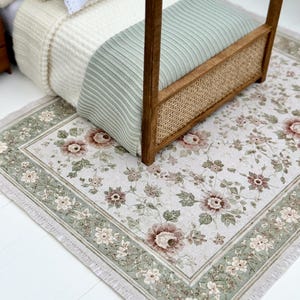Può includere: Un tappeto rettangolare con un motivo floreale di fiori rosa tenue e foglie verdi su sfondo crema, bordato da una cornice verde salvia con accenti floreali bianchi e frange, posizionato sotto un letto per casa delle bambole in legno.