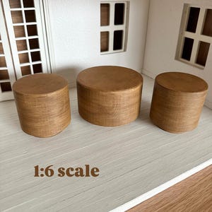 May include: Three miniature wooden cylinders, likely furniture, are arranged on a light-colored surface. The cylinders are a warm brown color and have a visible wood grain. The text "1:6 scale" is at the bottom of the image.