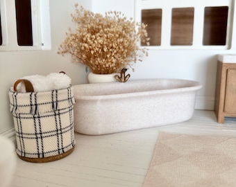 May include: A miniature bathroom scene featuring a light pink bathtub, a plaid basket, and a beige rug. A vase of dried flowers sits near the tub. The room has white walls and windows, creating a bright and airy atmosphere.