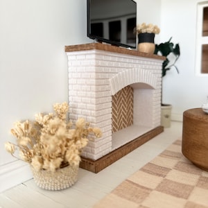 May include: A miniature white brick fireplace with a wooden mantel supports a black television. A woven basket holds dried flowers, and a checkered rug lies on the floor. The fireplace has a herringbone pattern inside the arch.
