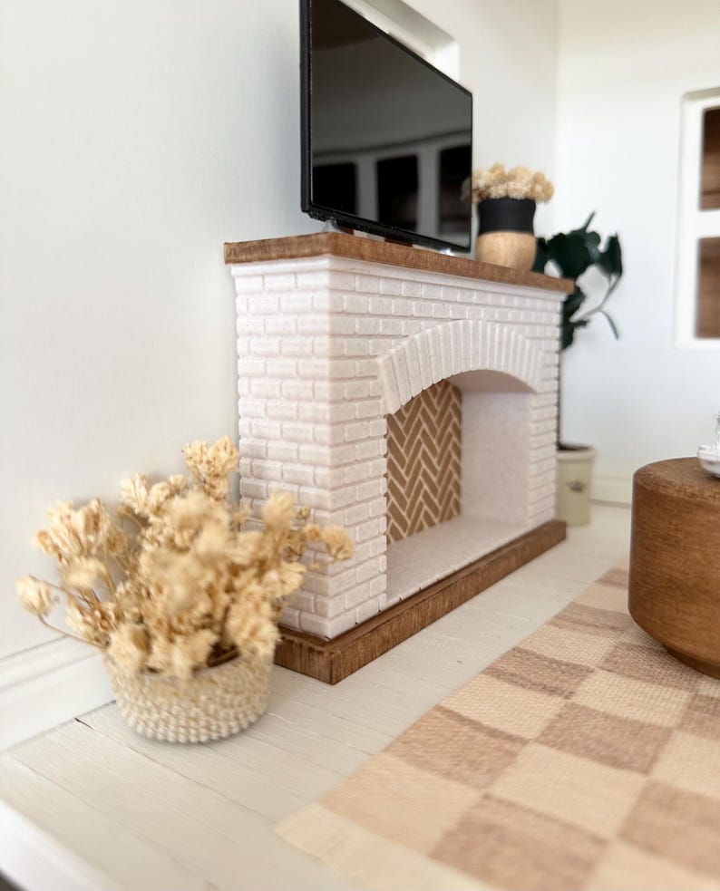 May include: A miniature white brick fireplace with a wooden mantel supports a black television. A woven basket holds dried flowers, and a checkered rug lies on the floor. The fireplace has a herringbone pattern inside the arch.