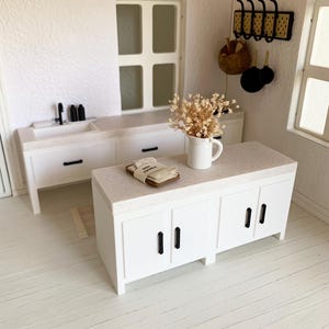 May include: White kitchen island with a white countertop and black cabinet hardware. The island has three drawers and two doors. A small bouquet of dried flowers is on the countertop.