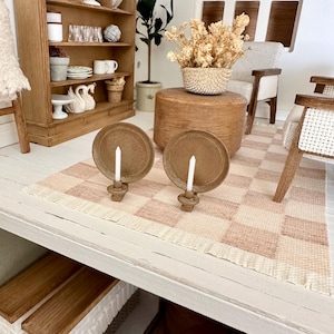 May include: Miniature dollhouse interior scene with two brown candle holders with white candles on a checkered rug. A wooden cabinet with decorative items and a small potted plant are in the background. A small basket of dried flowers sits on a wooden table.
