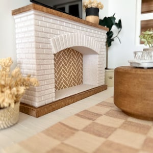 May include: Miniature fireplace with a white brick facade, arched opening, and a wooden mantel. The fireplace features a herringbone pattern inside the opening. A black television sits on top. A checkered rug and decorative plants complete the scene.