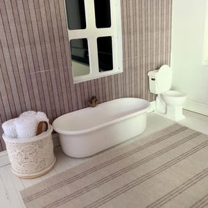 May include: A white bathtub with a white towel rack and a white toilet in a dollhouse bathroom with a striped rug and a window.