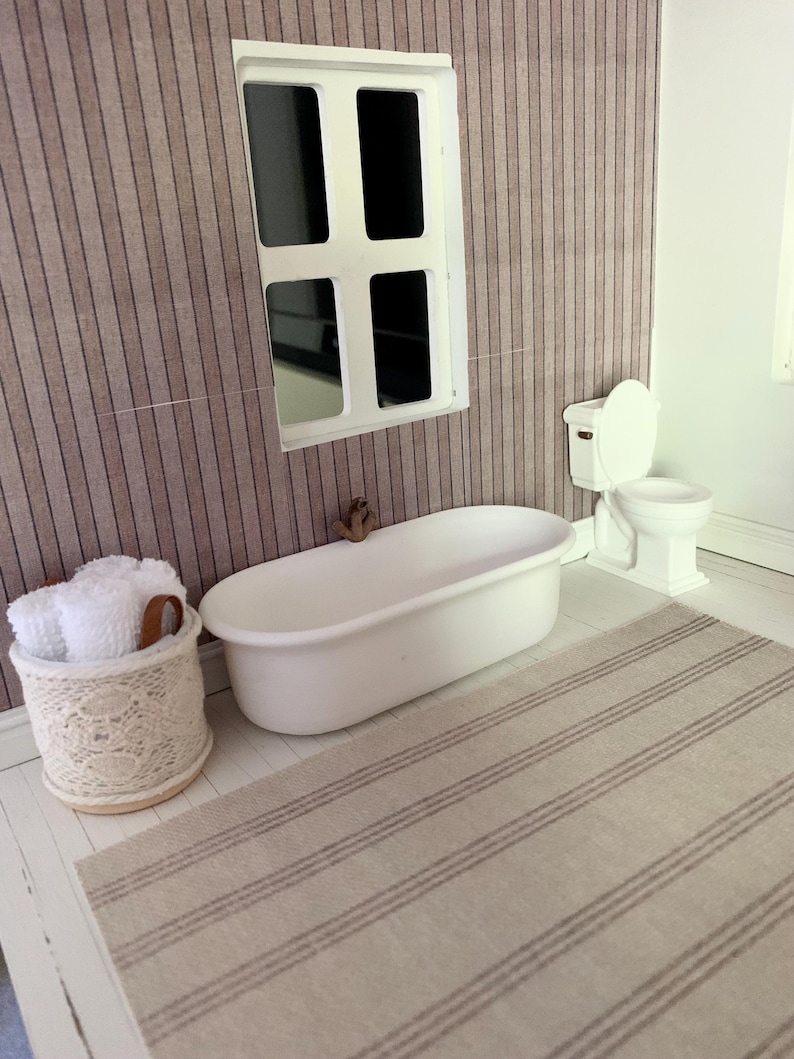 May include: A white bathtub with a white towel rack and a white toilet in a dollhouse bathroom with a striped rug and a window.