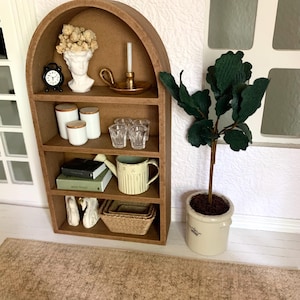 May include: A miniature wooden shelf unit with an arched top, featuring a variety of decorative items, including a bust, a candle holder, books, a watering can, and a potted plant. The shelf is set on a brown rug.