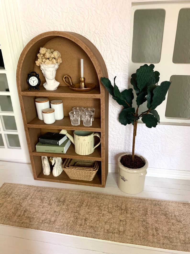 May include: A miniature wooden shelf unit with an arched top, featuring a variety of decorative items, including a bust, a candle holder, books, a watering can, and a potted plant. The shelf is set on a brown rug.