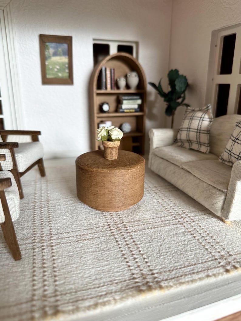 Op de afbeelding: Miniatuur woonkamersc&egrave;ne met een lichtgekleurde bank, stoelen en een ronde houten salontafel. Een kleine pot met witte bloemen staat op de tafel. Een boekenplank en ingelijste kunstwerken zijn zichtbaar op de achtergrond. Het tapijt heeft een ruitpatroon.