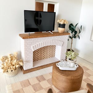 May include: Miniature living room scene featuring a white brick fireplace with a wooden mantel, a black flat-screen TV, and a small wooden ottoman. A checkered rug and potted plants complete the cozy decor.