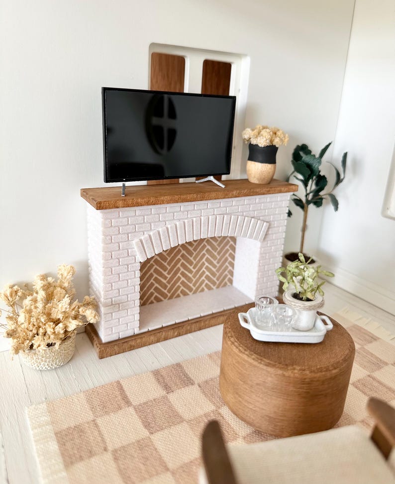May include: Miniature living room scene featuring a white brick fireplace with a wooden mantel, a black flat-screen TV, and a small wooden ottoman. A checkered rug and potted plants complete the cozy decor.