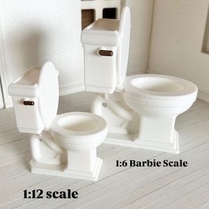 May include: Three white miniature toilets, with open and closed lids, are displayed. The toilets have brown accents and are labeled with "1:12 scale" and "1:6 Barbie Scale". The background is a light-colored, textured surface.