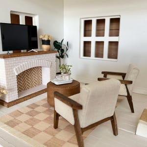 May include: Miniature living room scene with a white brick fireplace, a flat-screen TV, and two armchairs. A checkered rug and a small round table complete the decor. The room is painted white with a window.