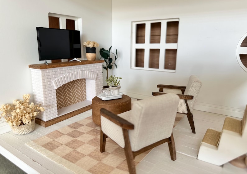 May include: Miniature living room scene with a white brick fireplace, a flat-screen TV, and two armchairs. A checkered rug and a small round table complete the decor. The room is painted white with a window.