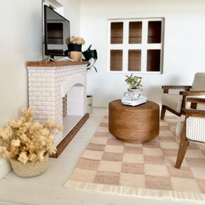 May include: Miniature living room scene with a white brick fireplace, a TV, and a brown wooden coffee table. A checkered rug in shades of beige and brown sits on the floor. The scene includes a window, a chair, and decorative plants.
