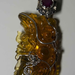 Puede incluir: Un colgante con una figura detallada de color ámbar con detalles de alambre plateado. El colgante tiene una parte superior plateada con una piedra preciosa roja. La figura tiene un rostro detallado y patrones arremolinados.