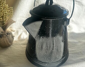 May include: Black enamel camping coffee pot with a speckled white finish and a wooden handle. The pot has a spout and a wire handle.