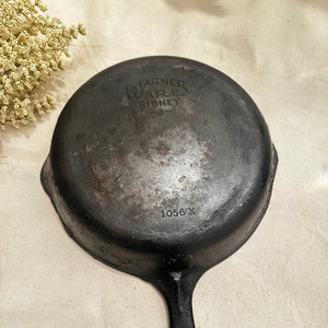 May include: A black cast iron skillet with a long handle and a circular cooking surface. The words "Wagner Ware Sidney -0-" and "1056 X" are embossed on the bottom of the pan. The skillet is vintage and shows signs of wear.