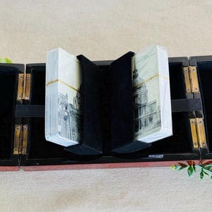 May include: A rectangular black wooden box with gold-toned metal hinges, featuring two open compartments lined with black velvet. Inside, two decks of playing cards with black and white architectural illustrations are held upright by black dividers.