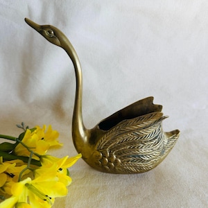 May include: A brass swan figurine with a long, curved neck and detailed wing feathers. The swan has a small opening on its back, possibly for holding small items. Yellow flowers are visible in the foreground.