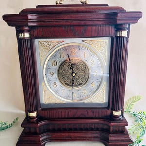 May include: A wooden mantel clock with a rectangular shape and a dark reddish-brown finish. The clock face is silver with gold accents and Roman numerals. The clock has a gold-colored handle on top and decorative columns on the sides.